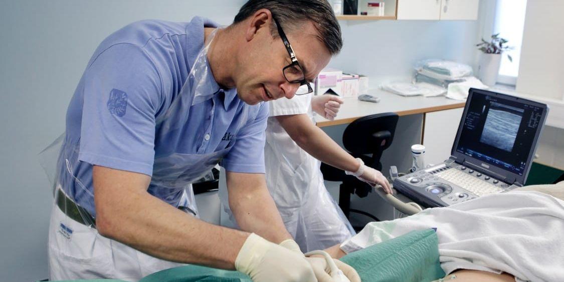 Peter Danielsson utför ingreppet i ett behandlingsrum på kirurgkliniken med hjälp av ultraljudsbild av blodådern på en bildskärm. Sjuksköterskan Linda Rapp skymtar i bakgrunden. 